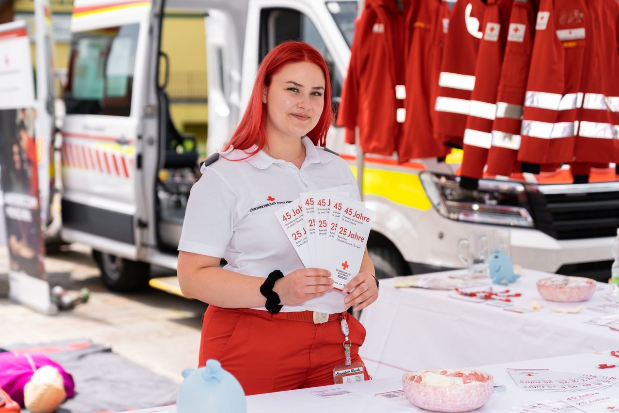 Eine Frau mit roten Haaren trägt ein weißes Uniform und hält Urkunden. Hinter ihr steht ein Krankenwagen mit offener Tür.
