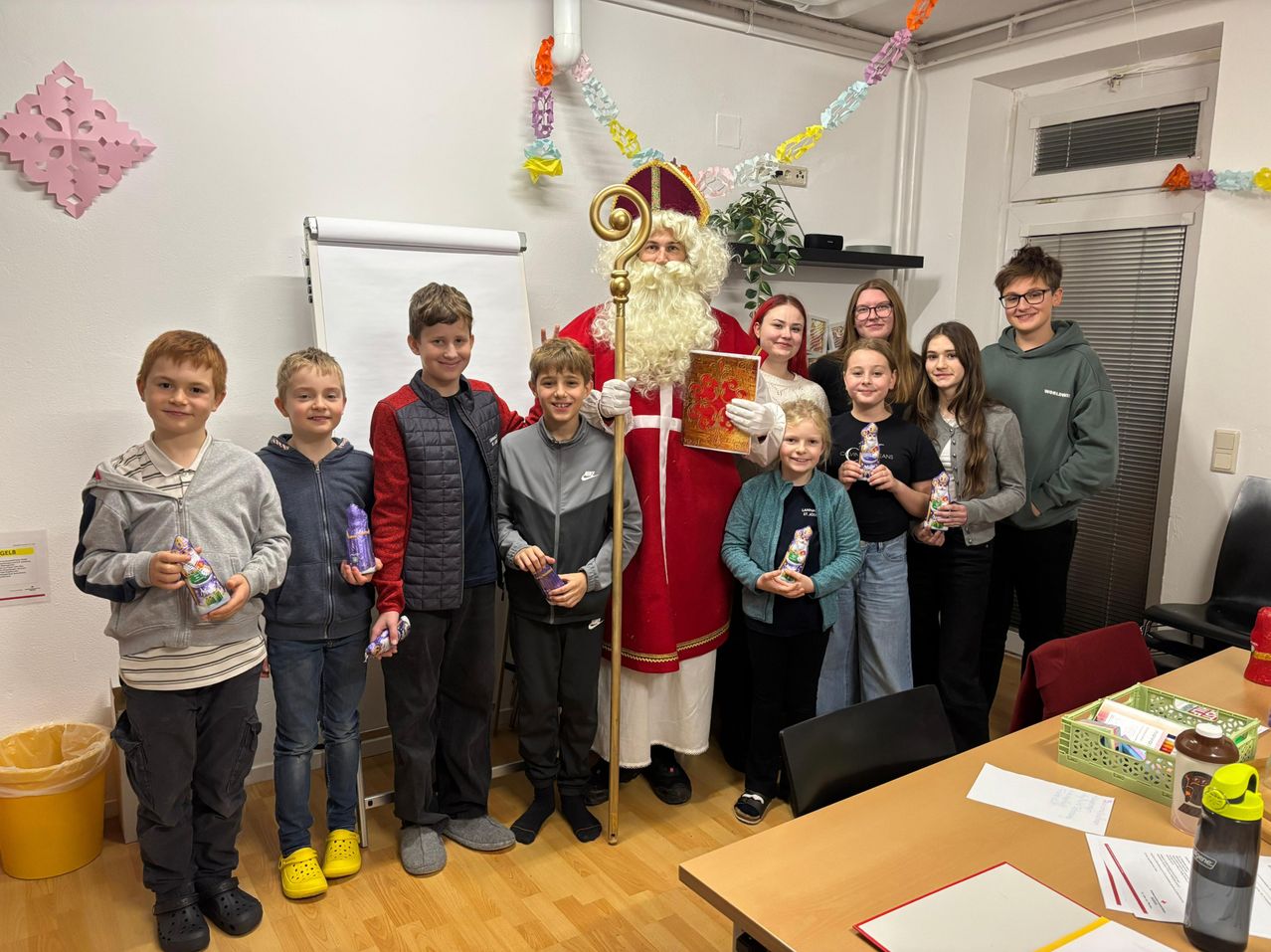 Eine Gruppe von Kindern und Erwachsenen posiert für ein Foto mit einem als Weihnachtsmann verkleideten Mann in einem Raum mit Holzböden und Dekorationen.