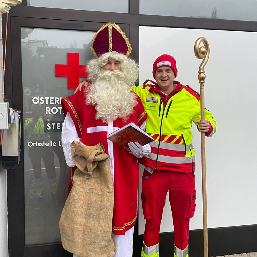 Ein Mann in der Kleidung des Heiligen Nikolaus hält ein Buch und einen Sack und steht neben einem Mann in einer reflektierenden Weste.