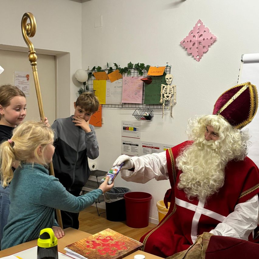 Eine Gruppe von Kindern versammelt sich um einen Mann, der als Weihnachtsmann verkleidet ist. Sie halten eine Box und ein Geschenk, während der Weihnachtsmann einen Stab und einen Handschuh hält. Der Schauplatz ist drinnen, mit einer weißen Wand und verschiedenen Dekorationen.