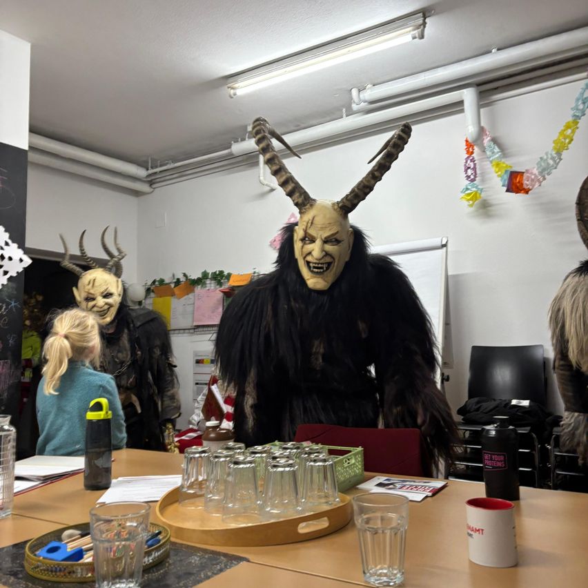 Ein Klassenzimmer mit Personen in Kostümen; eine Person in einem schwarzen Kostüm mit Hörnern steht an einem Tisch mit Gläsern, ein Mädchen sitzt an einem Schreibtisch und zwei weitere Personen in Kostümen stehen hinter dem Tisch.