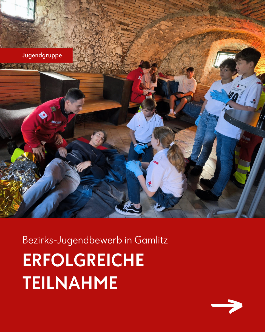 Eine Gruppe Jugendlicher in roten Jacken nimmt an einer Erste-Hilfe-Schulung teil, bei der eine verletzte Person auf einem Bett liegt. Ein Mädchen überprüft den Puls der verletzten Person.