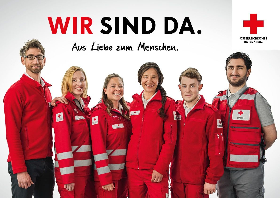 Eine Gruppe von Menschen in roten Uniformen mit dem Roten Kreuz Logo stehen zusammen und lächeln. Sie tragen alle eine Brille und eine Person trägt einen Anzug. Der Hintergrund ist weiß.