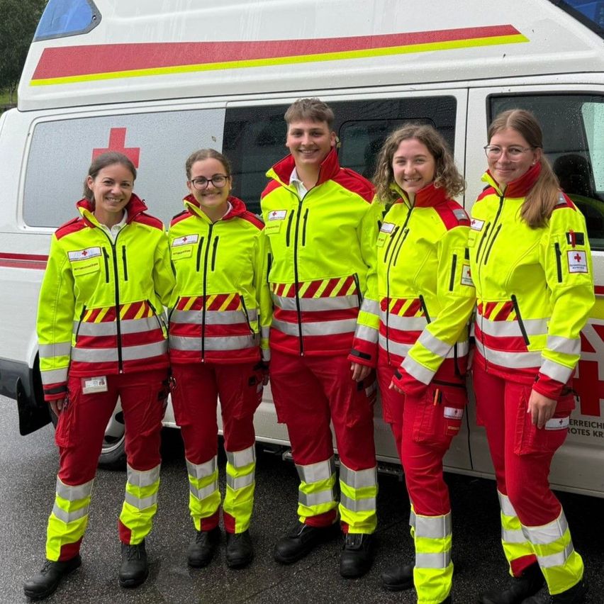 Fünf Personen in gelben und roten Uniformen stehen neben einem weißen Krankenwagen. Sie haben alle ernste Gesichtsausdrücke.