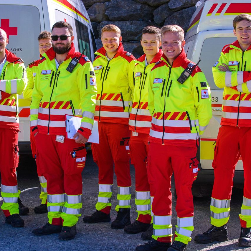 Sechs Personen in leuchtenden gelben und roten Sicherheitsanzügen stehen vor zwei Krankenwagen und lächeln für ein Foto.