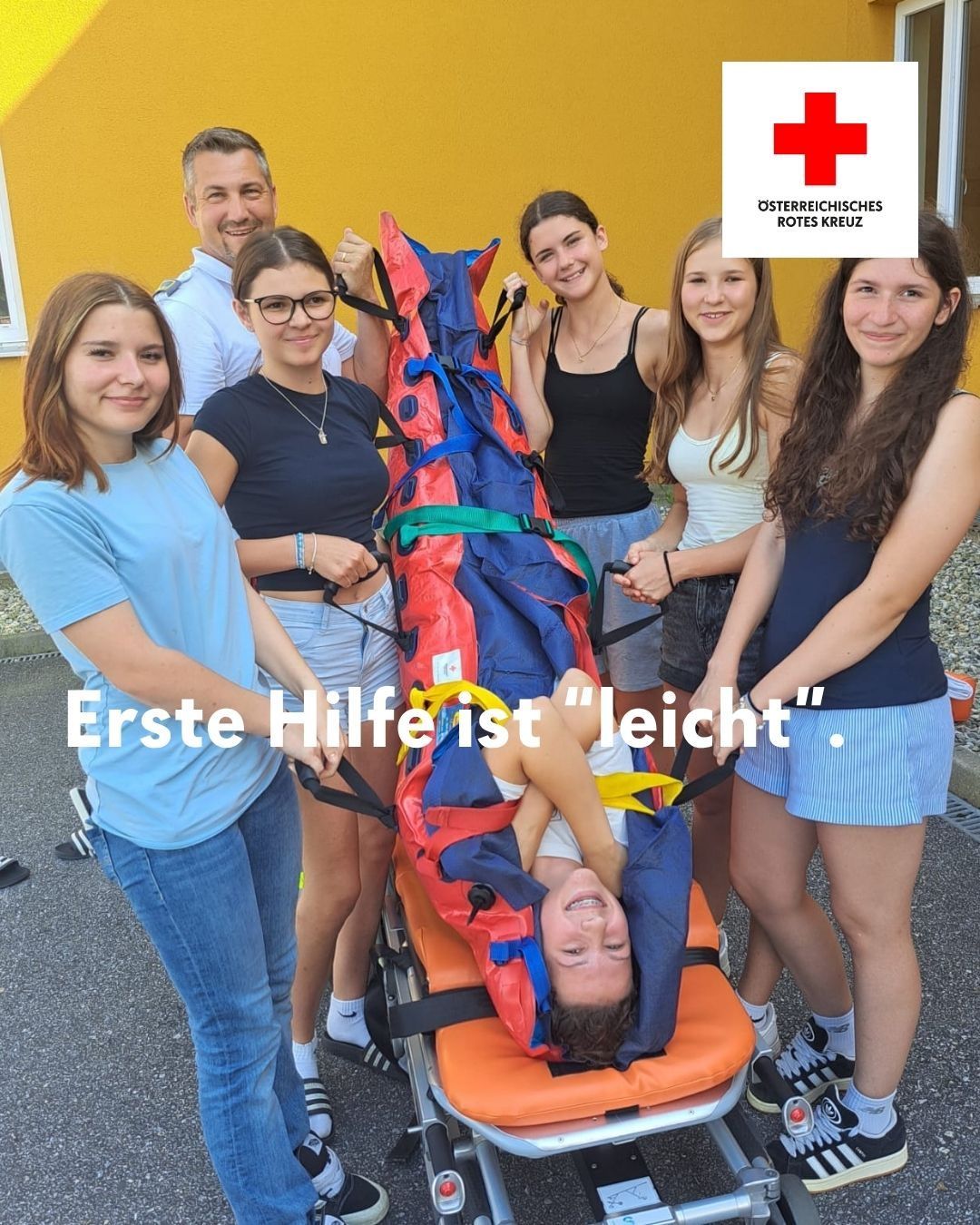 Eine Gruppe junger Frauen und Männer übt Erste Hilfe an einem Dummies, mit einem roten Kreuz im Hintergrund.