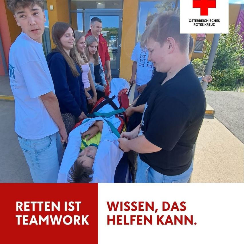 Eine Gruppe von Menschen, darunter junge Erwachsene, versammelt sich um eine Person auf einer Trage. Die Person auf der Trage wird von einer Person in einem schwarzen Shirt betreut. Die Szene spielt in einem medizinischen oder Notfallumfeld.