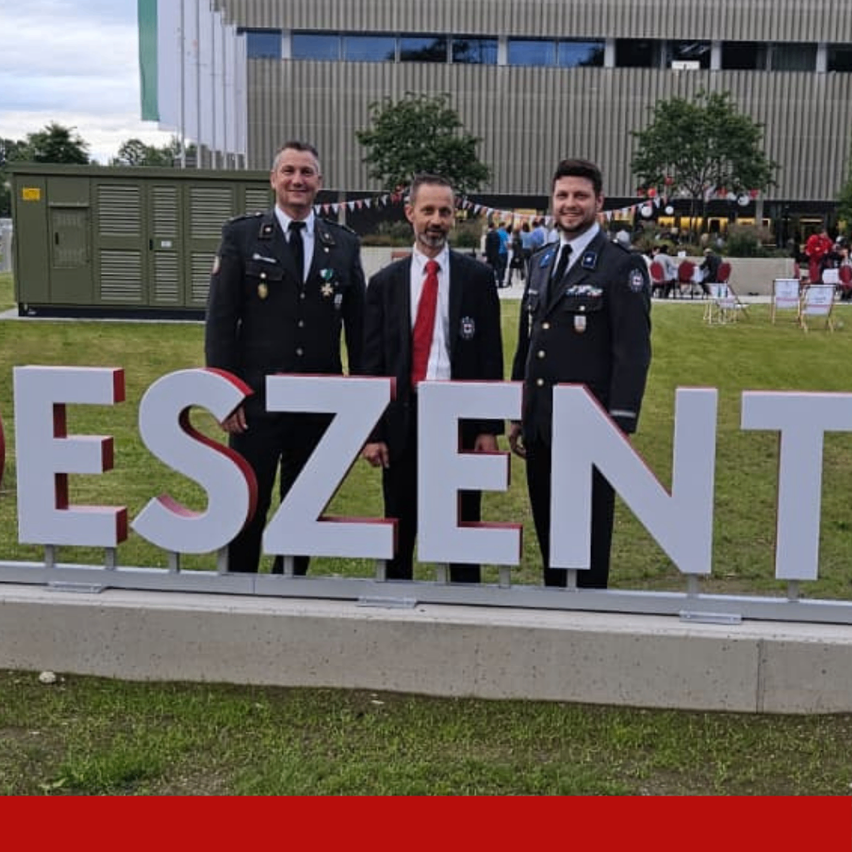 Drei Männer in Militäruniformen stehen vor einem großen Schild mit der Aufschrift ESZEN, mit einem Gebäude und Menschen im Hintergrund.