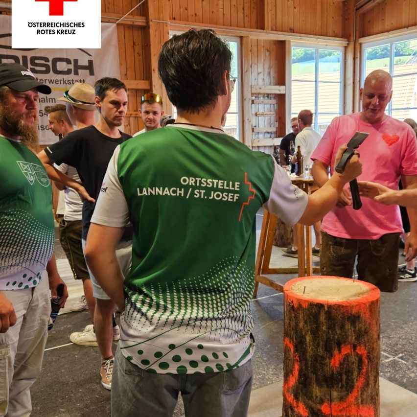 Mehrere Personen versammeln sich um einen Mann in einem grünen Shirt, der eine Axt hält, wahrscheinlich bei einem Wettbewerb. Der Mann steht vor einem Holzstumpf, und andere schauen zu. Die Umgebung scheint ein Werkstatt- oder Veranstaltungsraum zu sein.