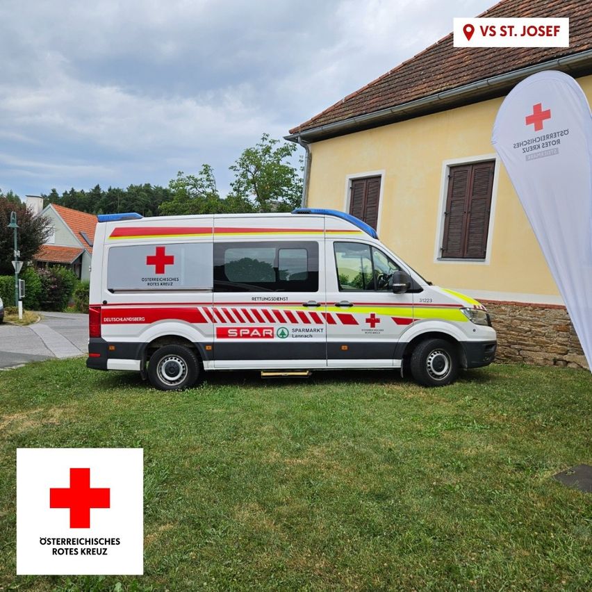 Ein Krankenwagen des Österreichischen Roten Kreuzes steht vor einem Gebäude mit einem roten Kreuzbanner.