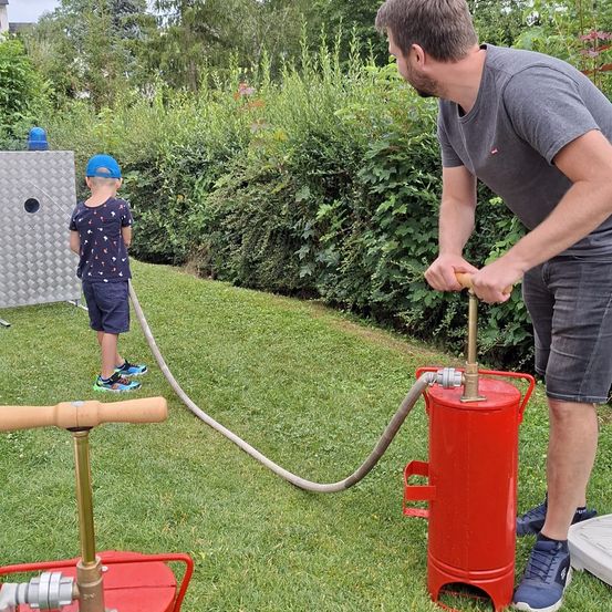 Ein Mann pumpt Wasser durch einen Schlauch in einen roten Behälter, während ein Kind zusieht. Der Rasen ist üppig und im Hintergrund befinden sich Büsche und Bäume.