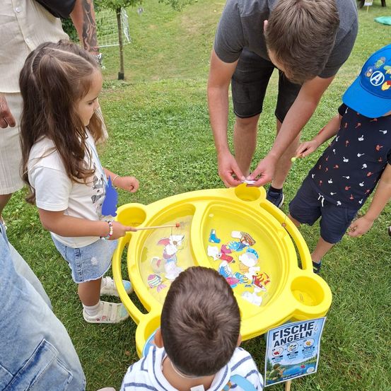 Ein Mann und Kinder sind draußen bei einem Angelspiel. Ein Kind hält eine Angelrute und der Mann hilft einem anderen Kind.