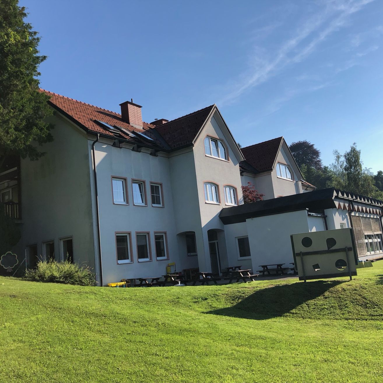 Ein großes Gebäude mit einem roten Dach und mehreren Fenstern. Solarpanele auf dem Dach. Picknicktische sind vor dem Gebäude aufgestellt.