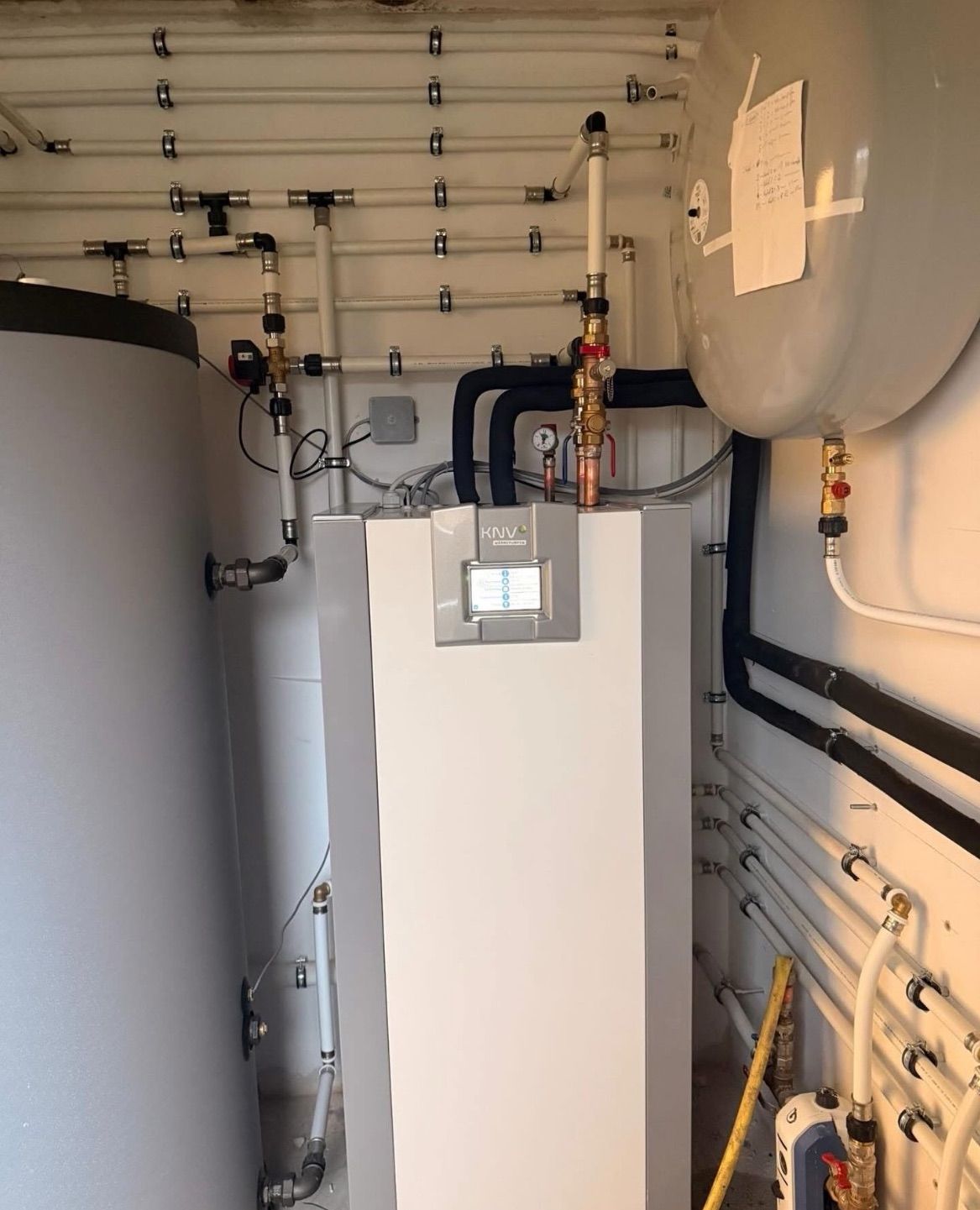 Ein Innenbereich mit weißen und schwarzen Rohren, einem zentralen KNV-Wassererhitzer und zwei großen Wassertanks.