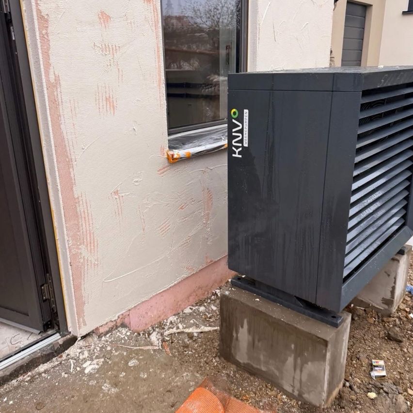 Ein schwarzes KNVO-Gerät steht auf einem Betonblock vor einem Gebäude. Die Wand hat einen rosa Rand und ein Fenster mit einer klaren Glassicht auf Bäume.