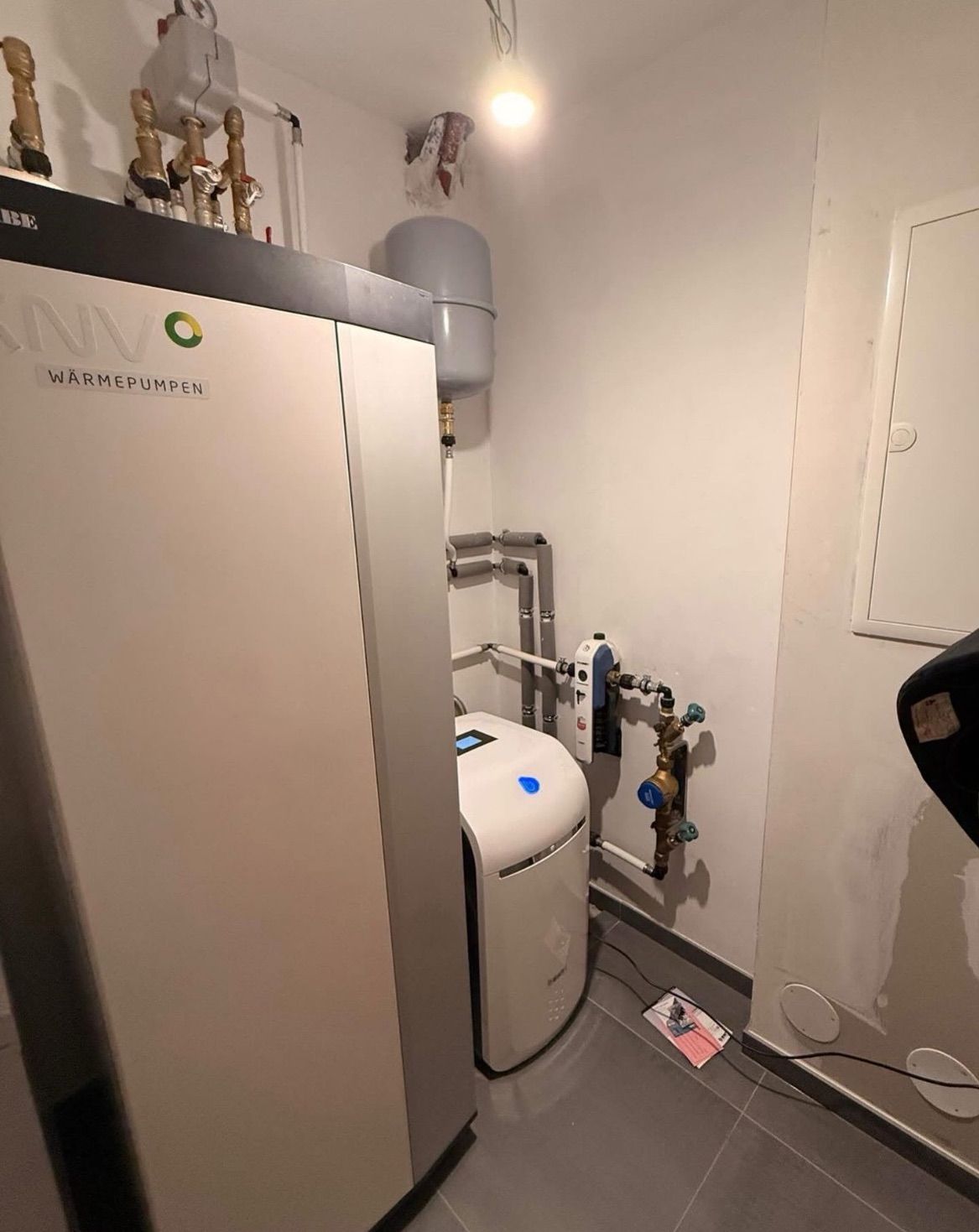 Eine weiße Wärmepumpe mit der Aufschrift ANVO ist an einer Wand montiert, mit einem Wassertank und Rohren. Ein kleiner Monitor befindet sich auf der Vorderseite der Pumpe.