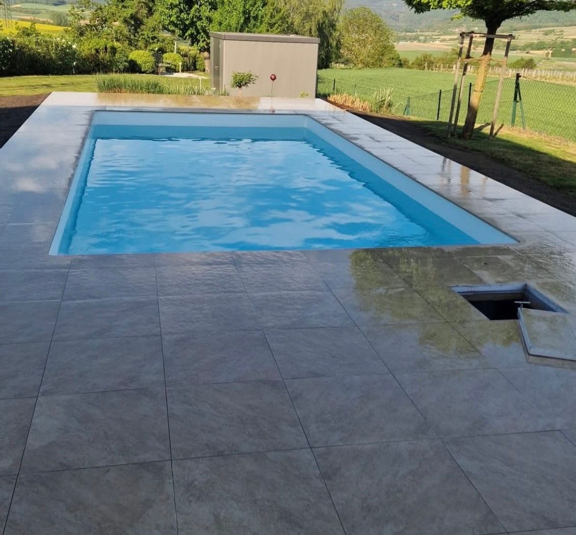 Ein leerer rechteckiger Swimmingpool mit gefliestem Umfeld. Ein kleines Gebäude und Bäume im Hintergrund.