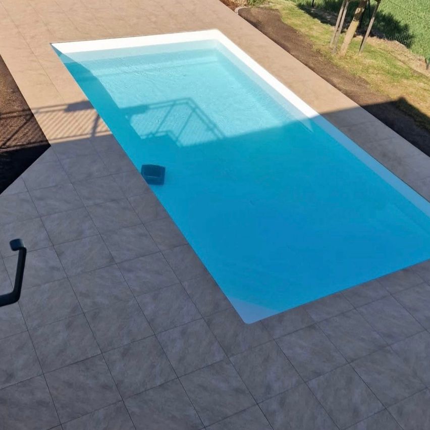 Eine Draufsicht auf einen rechteckigen Swimmingpool mit klarem blauem Wasser, umgeben von gefliesten Böden und einem Zaun auf der rechten Seite.