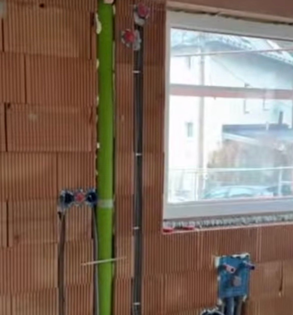A partially constructed interior wall with brown panels and a green pipe. A window with a view of a building and a car is visible.