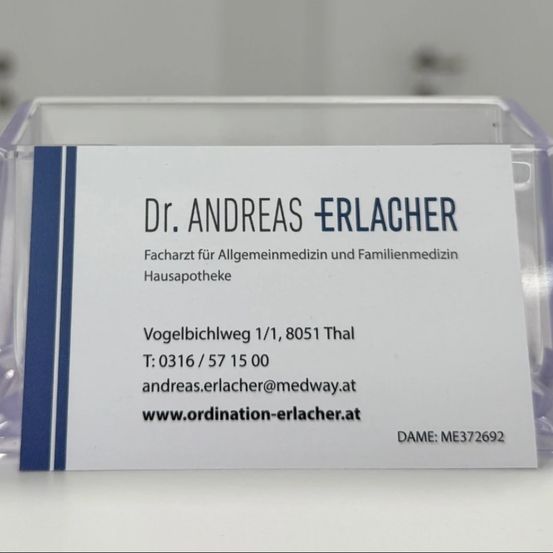 Ein durchsichtiger Kunststoffhalter zeigt eine weiße Visitenkarte mit blauen Akzenten. Die Karte trägt den Namen 'Dr. Andreas Erlacher', Facharzt für Allgemeinmedizin und Familienmedizin, mit Kontaktdaten wie Adresse, Telefonnummer, E-Mail und Website.