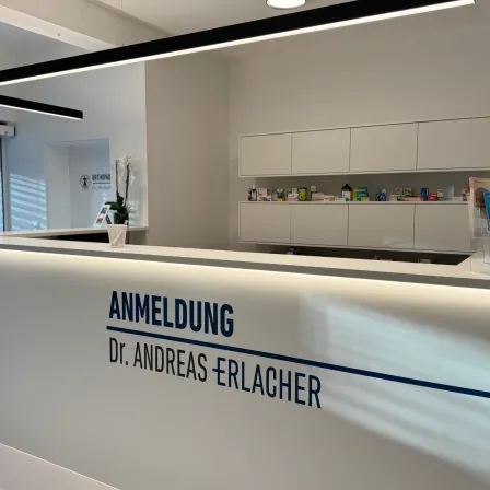 Eine weiße Empfangstheke mit blauem Text, der 'ANMELDUNG Dr. Andreas Erlacher' lautet. Dahinter befinden sich Regale mit medizinischen Utensilien und ein Logo an der linken Wand.