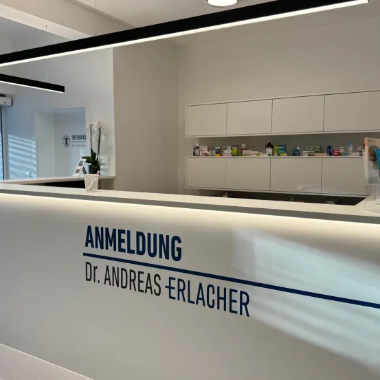 Eine weiße Empfangstheke mit blauem Text, der 'ANMELDUNG Dr. Andreas Erlacher' lautet. Dahinter befinden sich Regale mit medizinischen Utensilien und ein Logo an der linken Wand.