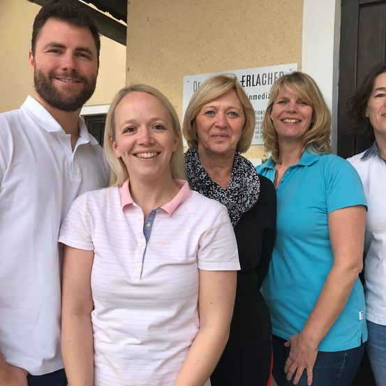 Fünf Menschen stehen lächelnd für ein Foto vor einem Gebäude. Die Frau in der Mitte trägt ein Halstuch, während die anderen Freizeitkleidung tragen. Hinter ihnen befindet sich ein Schild mit Text.