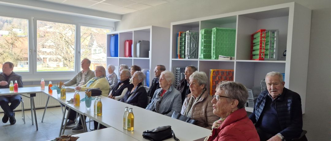 Eine Gruppe älterer Menschen sitzt in einem Raum mit weißen Wänden, Regalen mit verschiedenen Gegenständen und einem Fenster, das einen Blick nach draußen ermöglicht. Sie sitzen vorn und sehen nach vorne, mit Flaschen und einer Handtasche auf einem Tisch vor ihnen.