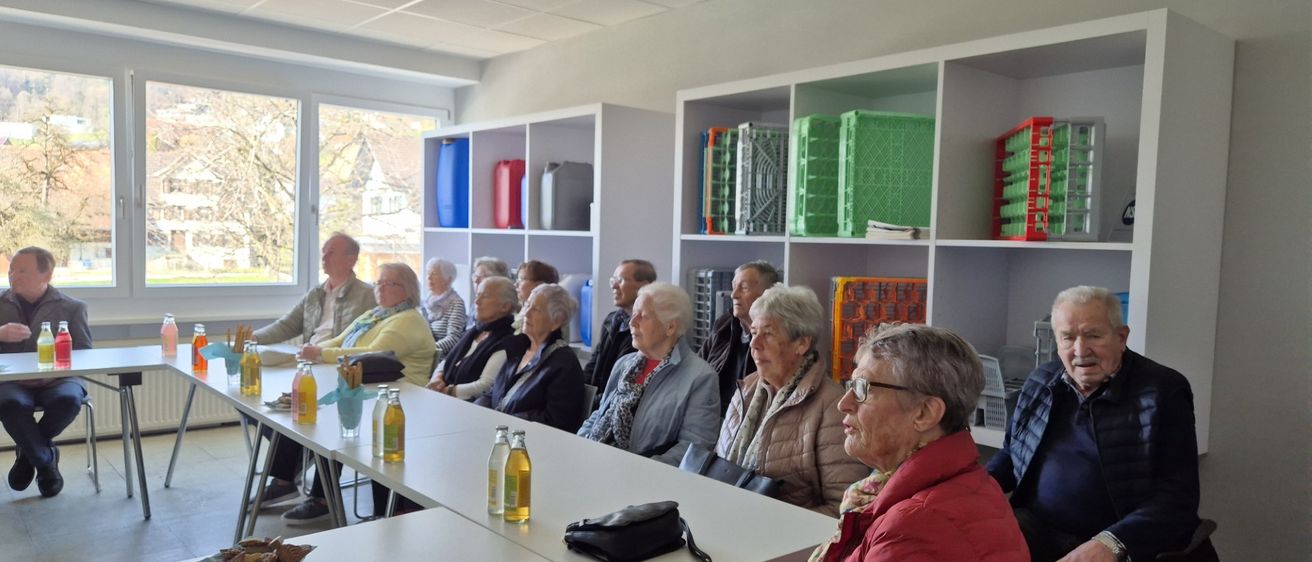 Eine Gruppe älterer Menschen sitzt in einem Raum mit weißen Wänden, Regalen mit verschiedenen Gegenständen und einem Fenster, das einen Blick nach draußen ermöglicht. Sie sitzen vorn und sehen nach vorne, mit Flaschen und einer Handtasche auf einem Tisch vor ihnen.