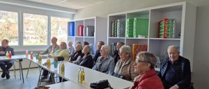 Eine Gruppe älterer Menschen sitzt in einem Raum mit weißen Wänden, Regalen mit verschiedenen Gegenständen und einem Fenster, das einen Blick nach draußen ermöglicht. Sie sitzen vorn und sehen nach vorne, mit Flaschen und einer Handtasche auf einem Tisch vor ihnen.