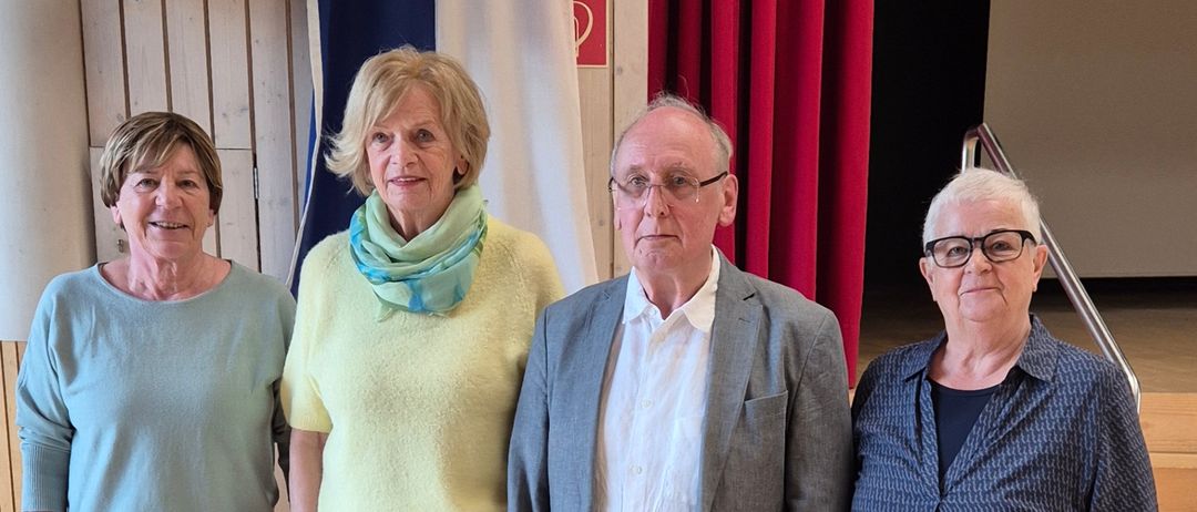 Vier Erwachsene stehen lächelnd für die Kamera auf einem Holzboden. Zwei Frauen und ein Mann tragen Brillen. Hinter ihnen ist ein roter Vorhang und eine Flagge.