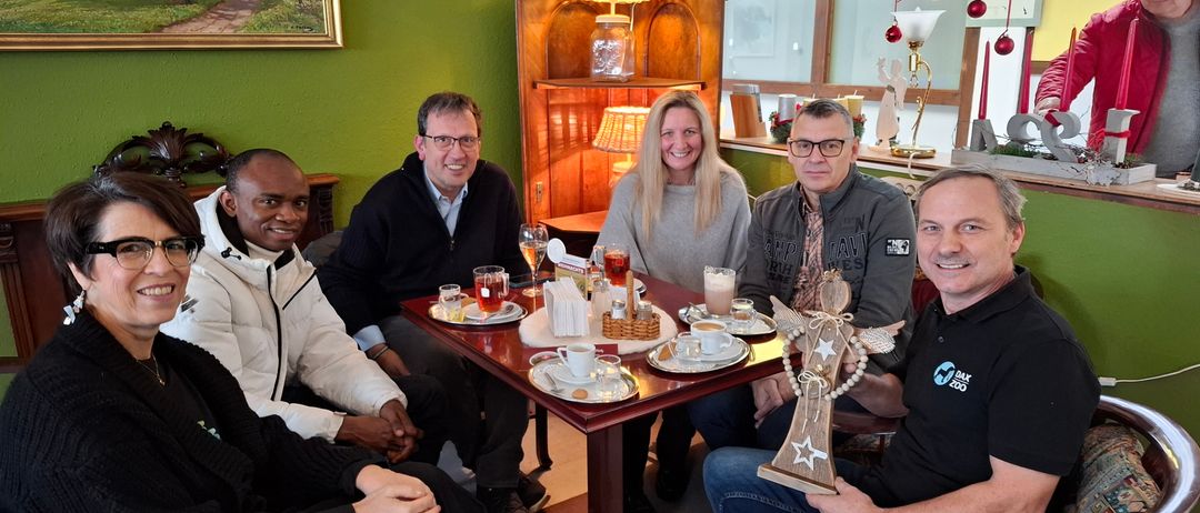 Eine Gruppe von Menschen sitzt in einem Café um einen Tisch herum und lächelt. Der Tisch hat verschiedene Getränke und Snacks. Ein Mann auf der rechten Seite hält einen hölzernen Engel. Hinter ihnen ist ein Schrank mit Gläsern und einer Lampe zu sehen.