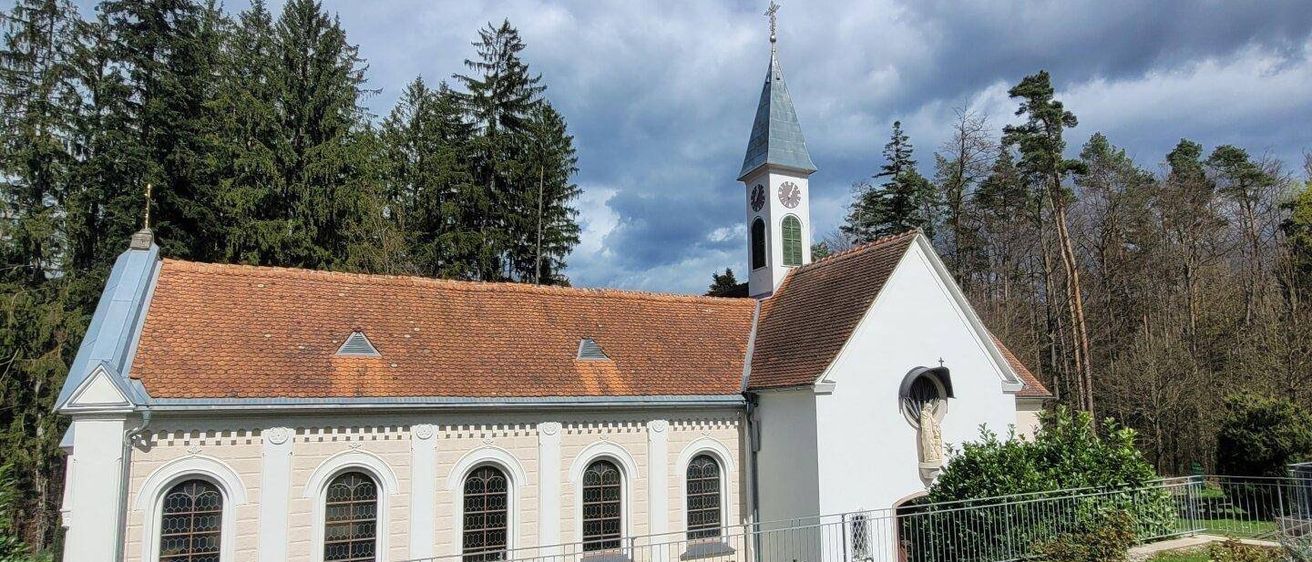 Eine weiße Kirche mit einem braunen Dach und einem Turm mit einer Uhr steht umgeben von Bäumen. Sie hat Bogenfenster und eine Statue an der Seite, umgeben von Grün und einem Metallzaun.