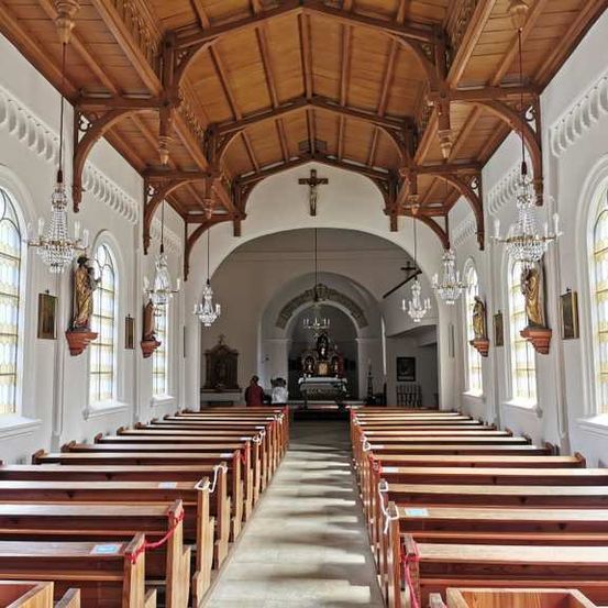 Das Innere einer Kirche mit Holzbänken, einer Holzdecke und Buntglasfenstern. Altäre und Statuen sind vorhanden, und zwei Personen befinden sich in der Nähe des Altars.