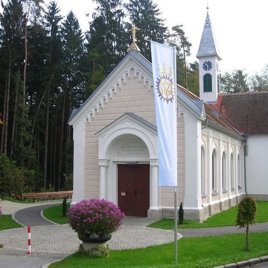Eine kleine weiße Kirche mit einer braunen Tür und einem Glockenturm. Ein Banner ist an einem Pfosten vor der Kirche befestigt. Ein Weg führt zum Eingang.