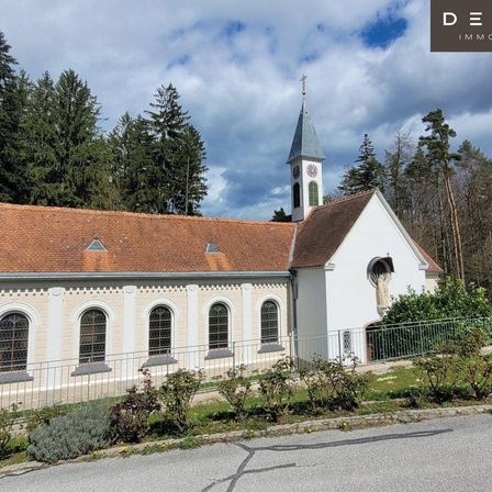 Eine kleine weiße Kirche mit braunem Dach und einem Glockenturm steht in einem bewaldeten Gebiet. An der Seite der Kirche befindet sich eine Statue. Das Gebäude ist von einem Metallzaun und Grünflächen umgeben.