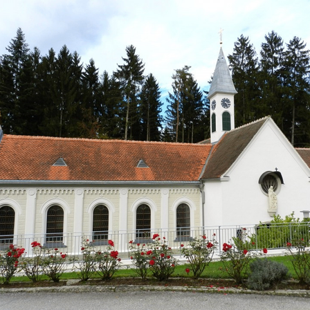 Eine weiße Kirche mit einem braunen Dach und einem Turm mit einer Uhr. Sie ist von roten Blumen und einem Zaun umgeben. Bäume sind im Hintergrund.