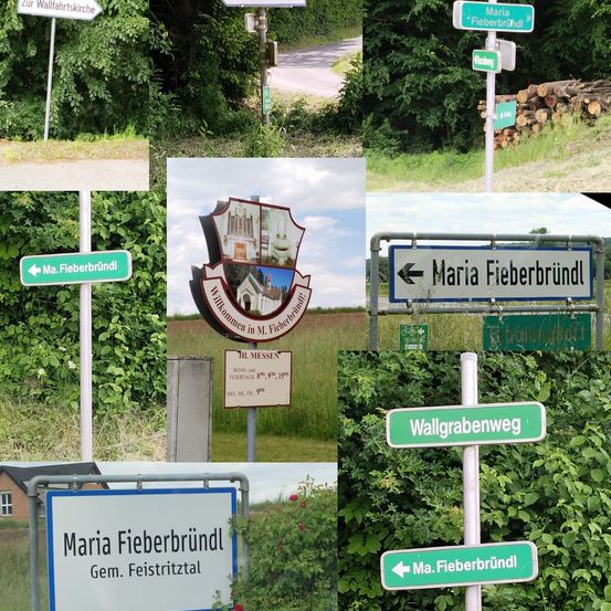 Mehrere Straßenschilder in einem ländlichen Gebiet. Ein Schild weist auf Maria Fieberbründl hin, ein anderes auf Wallgrabenweg. Ein drittes Schild ist weiß mit blauem Text.
