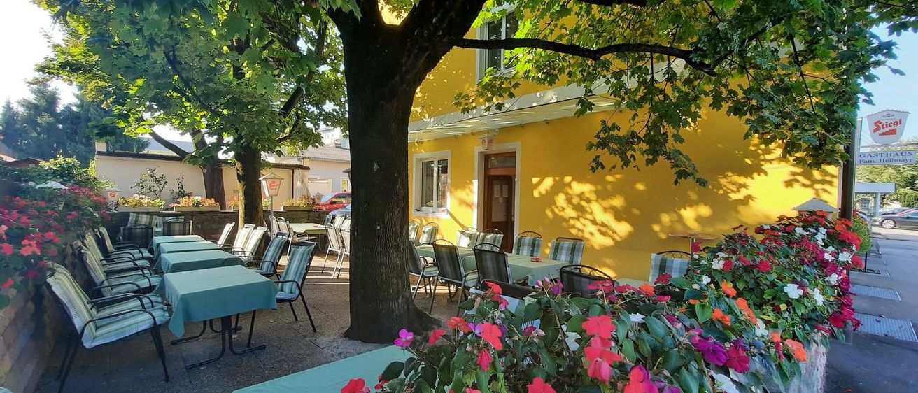 Eine Außensitzfläche eines Restaurants mit gelben Wänden, Bäumen und Blumen. Tische und Stühle sind für Gäste vorbereitet.