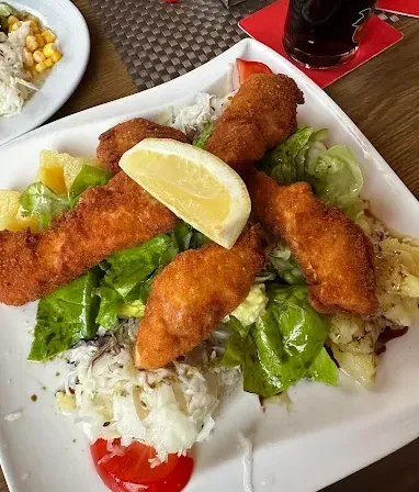 Ein weißer Teller mit gebratenen Fischfilets, Salat, einer Zitronenscheibe und einer Tomate. Ein Glas Limonade und ein weiterer Teller im Hintergrund.