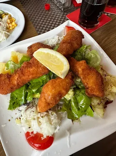 Ein weißer Teller mit gebratenen Fischfilets, Salat, einer Zitronenscheibe und einer Tomate. Ein Glas Limonade und ein weiterer Teller im Hintergrund.