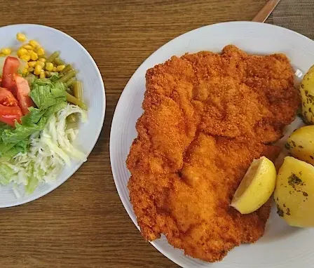 Zwei Teller auf einem Holztisch. Einer mit paniertem Schweinekotelett, Kartoffeln und Zitrone. Der andere mit Salat, Tomaten, Mais und Bohnen.