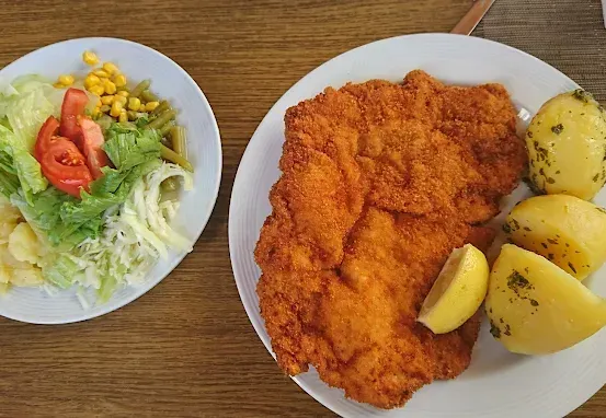 Zwei Teller auf einem Holztisch. Einer mit paniertem Schweinekotelett, Kartoffeln und Zitrone. Der andere mit Salat, Tomaten, Mais und Bohnen.