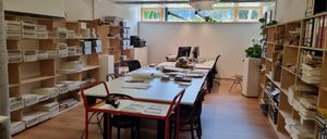 Ein Büro mit zwei langen Tischen, Computern, Büchern und Regalen. Es gibt auch Stühle, einen Schreibtisch und einen Drucker. Der Raum hat Fenster mit Pflanzen draußen.