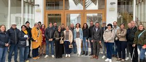A group of people dressed in winter attire stands outside a modern building with glass doors and a wooden frame.