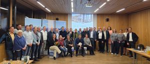Eine Gruppe von Menschen posiert für ein Foto in einem Konferenzraum mit einer Projektionsleinwand, die eine Präsentation zeigt.
