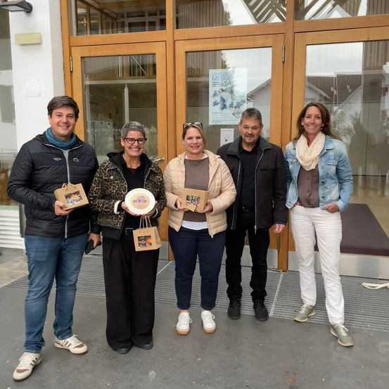 Fünf Personen stehen vor einem Gebäude. Sie alle haben Lächeln im Gesicht und halten Geschenke in den Händen.