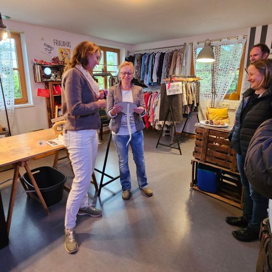 Fünf Frauen sind in einem Raum mit Kleiderständern, einem Tisch und einem Holzkasten. Zwei Frauen halten Papiere und zwei Frauen schauen sie an.