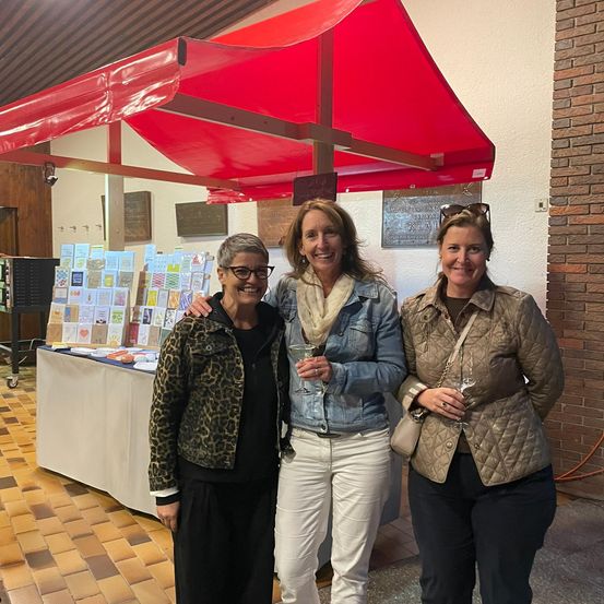 Drei Frauen stehen unter einem roten Vordach, lächeln für ein Foto. Sie halten Weingläser in der Hand und stehen vor einem Tisch mit Karten und einer Backsteinmauer.