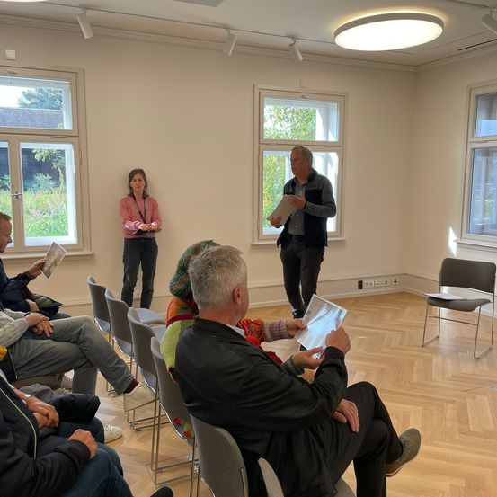 In einem Raum mit weißen Wänden sitzen mehrere Personen in Stühlen, die einem Mann mit Papieren zugewandt sind. Der Mann scheint eine Präsentation zu halten, während zwei weitere hinter ihm stehen, darunter eine Frau. Der Raum hat große Fenster und einen Holzboden.